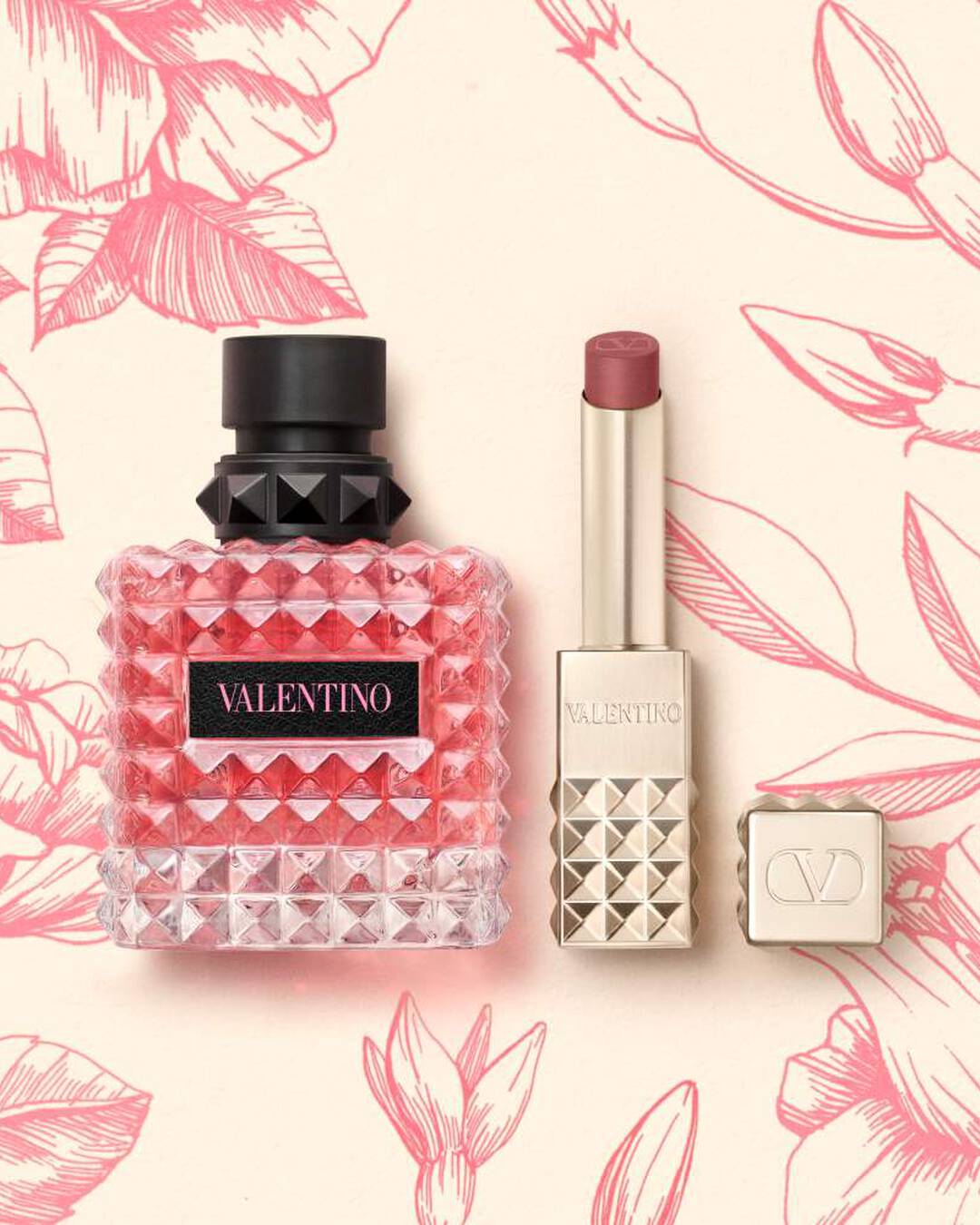 A pink Valentino perfume bottle and a lipstick in a square tube with stud detailing, set against a cream background with pink floral illustrations.