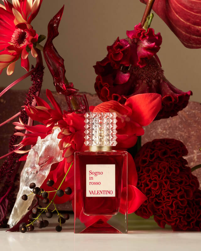 Una bottiglia di profumo Valentino circondata da fiori rossi e decorazioni floreali, su uno sfondo neutro.