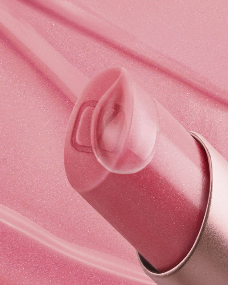 Un primo piano di un balsamo labbra rosa, con il suo tappo trasparente e traslucido leggermente aperto, che rivela un sottile logo "V" all'interno, il tutto su uno sfondo rosa tenue con leggere striature di prodotto.
