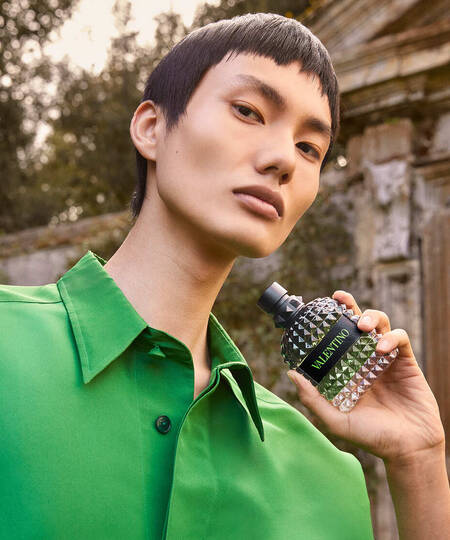 Giovane modello con camicia verde, tiene in mano un profumo Valentino in un contesto naturale e raffinato.