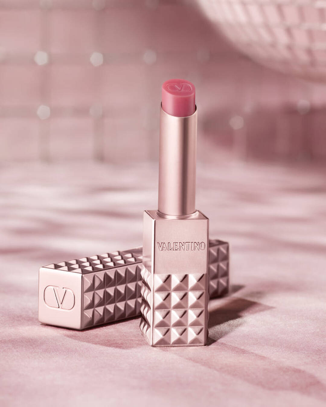 Un prodotto per labbra Valentino Spike color oro rosa con la punta rosa e in posizione verticale, con la sua custodia borchiata abbinata distesa accanto. Entrambi gli articoli sono posizionati su uno sfondo rosa tenue e scintillante che evoca un senso di glamour.
