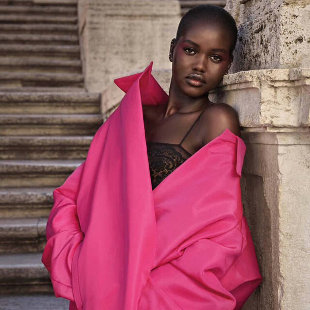Modella con pelle scura, vestita con un grande cappotto rosa, posa su scale di pietra, esprimendo eleganza e stile.