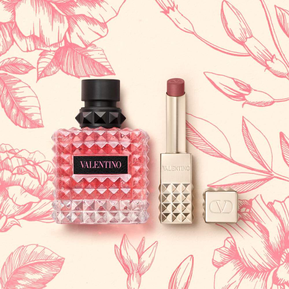 A pink Valentino perfume bottle and a lipstick in a square tube with stud detailing, set against a cream background with pink floral illustrations.
