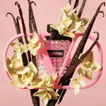 A pink Valentino perfume bottle is elegantly displayed within a stylish pink frame, surrounded by vanilla pods and delicate yellow flowers against a soft pink background.