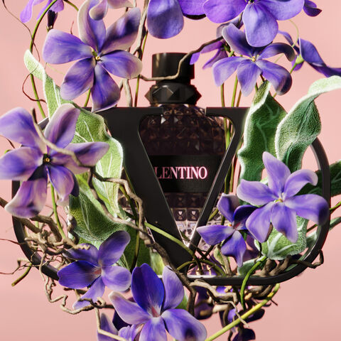 A bottle of Valentino perfume surrounded by vibrant purple flowers and lush greenery, set against a soft pink background.