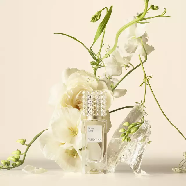 A Valentino perfume bottle arranged with white flowers and translucent crystals on a neutral background.