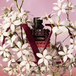 A stylish perfume bottle surrounded by white flowers and framed by a stylized "V" shape, set against a soft pink background.