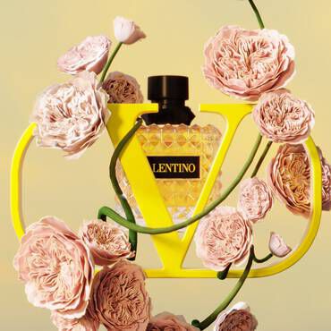 A vibrant yellow perfume bottle surrounded by delicate pink flowers, framed by a bold yellow "V" logo.