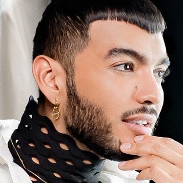 Un uomo con capelli corti e barbetta, che indossa una camicia bianca e una sciarpa nera a pois, guarda pensieroso verso uno specchio, con luci brillanti sullo sfondo.