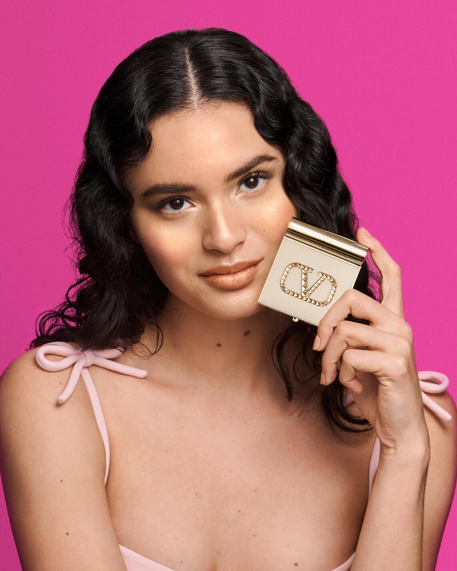 A woman with wavy hair poses against a bright pink background, holding a stylish compact. She is wearing a light pink top and has a subtle glam look.