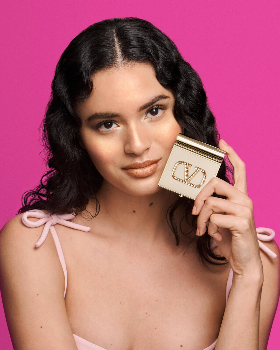 A woman with wavy hair poses against a bright pink background, holding a stylish compact. She is wearing a light pink top and has a subtle glam look.