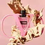A stylish pink perfume bottle enclosed in a large, sculptural letter "V" against a soft pink background, surrounded by organic, textured wood elements.