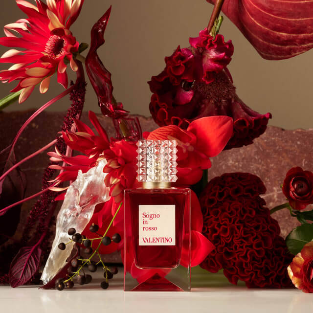 A bottle of Valentino perfume labeled "Sogno in Rosso" surrounded by vibrant red flowers and rich maroon textures, creating an elegant and luxurious aesthetic.