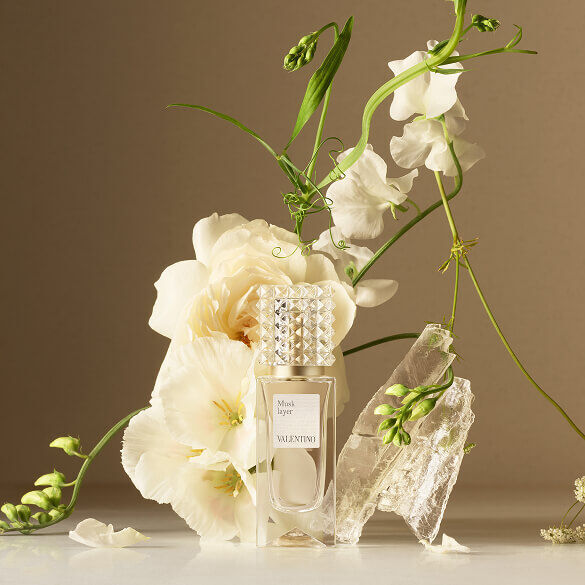 Un flacone di profumo Valentino disposto con fiori bianchi e cristalli traslucidi su uno sfondo neutro.
