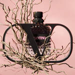 A stylized perfume bottle with a diamond-patterned design is framed by a black, abstract structure, surrounded by twisted, bare branches against a soft pink background. The label reads "Valentino."