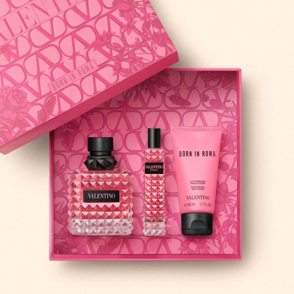 A pink gift box containing three Valentino beauty products: a large perfume bottle, a travel-sized perfume, and a scented lotion, all adorned with a diamond-patterned design.