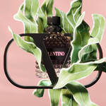A stylish perfume bottle with a black diamond-patterned design is surrounded by vibrant green leaves, all set against a soft pink background. The bottle features the brand name "Valentino" prominently displayed.