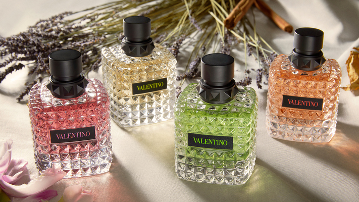 Quattro profumi Valentino in eleganti flaconi colorati su un tavolo, circondati da fiori e erbe aromatiche.